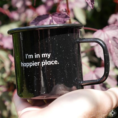 Black speckled "I'm in my happier place." enamel metal mug in its natural environment: outdoors.