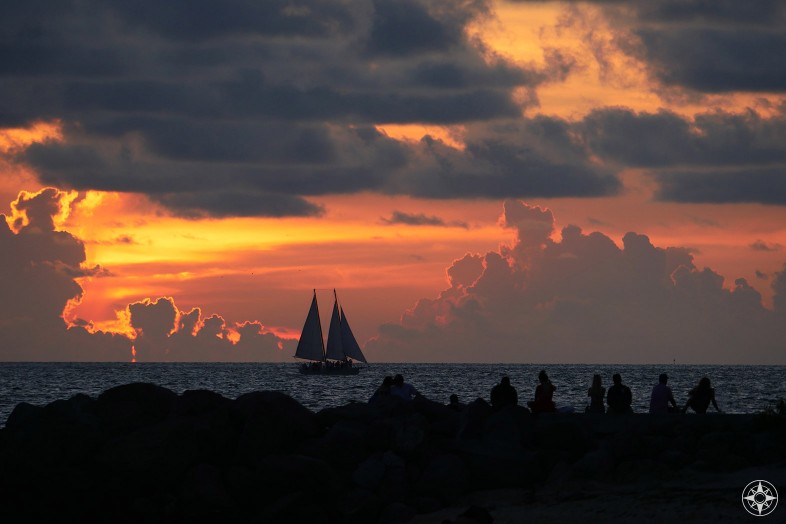 Sunset with silhouetted sailboat and people watching from rock wall