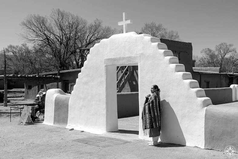 Native American woman, Taos Pueblo church, woven blanket, white wall gate with cross, merchant man, New Mexico, black and white
